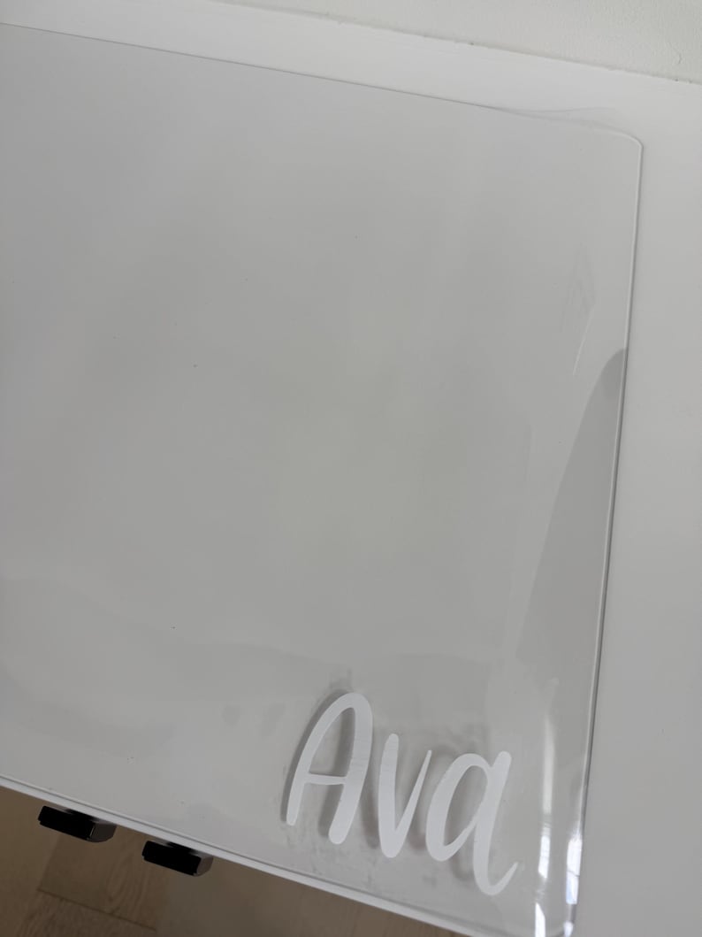 May include: A clear, rectangular acrylic sheet featuring the name Ava in white, cursive-style lettering applied to the surface. The transparent material has smooth, rounded corners and a glossy finish, reflecting light across its flat, clean surface.
