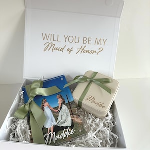 May include: A white gift box with the text "WILL YOU BE MY Maid of Honor?" printed inside the lid. The box contains a photo frame featuring a picture of two people and the name "Maddie," and a beige square jewelry box labeled "Maddie."
