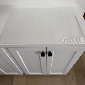 May include: A white cabinet with two doors featuring black square knobs. A clear, rectangular protective mat covers the top surface of the cabinet. The name Averie is written in white script lettering on the bottom right corner of the mat.