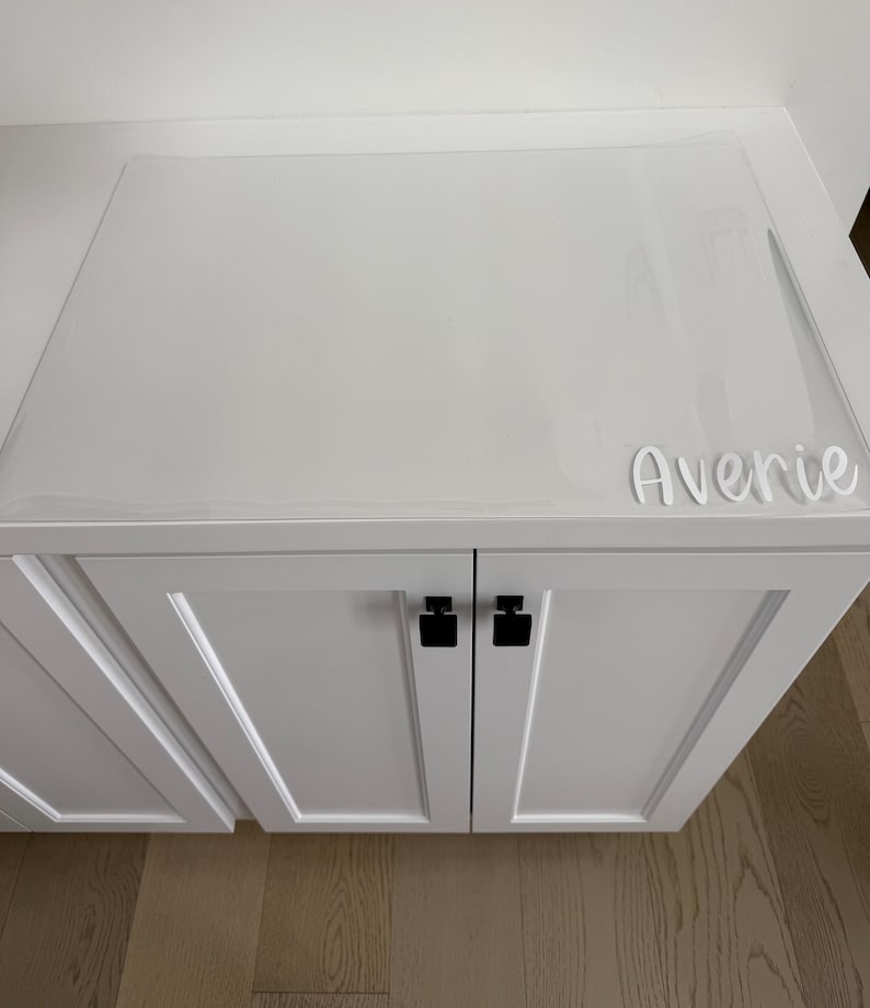 May include: A white cabinet with two doors featuring black square knobs. A clear, rectangular protective mat covers the top surface of the cabinet. The name Averie is written in white script lettering on the bottom right corner of the mat.