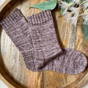 Könnte beinhalten: Ein Paar handgestrickte Socken in Burgunder-, Malven- und gesprenkelten Blau- und Violetttönen. Die Socken werden auf einem Holztablett präsentiert, das ihr strukturiertes Strickmuster und die gerippten Bündchen zeigt. Ein gemütliches Accessoire.