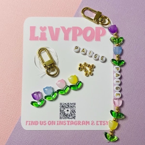 May include: A personalized keychain with the name "PAIGE" spelled out in white beads. The keychain features colorful tulip-shaped charms and a gold-colored clasp. The card behind the keychain has the text "LIVYPOP" and "FIND US ON INSTAGRAM & ETSY".
