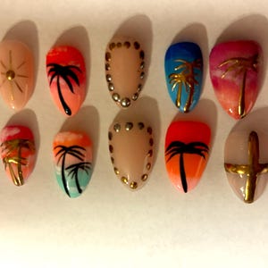 Könnte beinhalten: Ein Set aus zehn künstlichen Fingernägeln mit verschiedenen Designs, darunter Palmensilhouetten, Sonnenuntergangsverläufe in Orange, Pink und Blau, goldene Metallic-Akzente und kleine Metallnieten auf einer hellen, neutralen Basis.