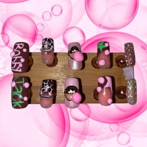 May include: A collection of ten artificial nails with diverse designs. The nails showcase pink, brown, and green hues, incorporating cartoon characters, text, and decorative features. The nails are presented on a wooden surface, set against a pink bubble backdrop.