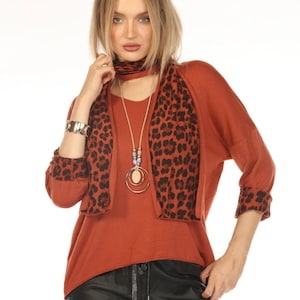 Puede incluir: Top color óxido con bufanda a juego con estampado de leopardo y pulsera. El conjunto incluye un collar largo con un colgante circular y pantalones de cuero negros. Las mangas del top tienen puños con estampado de leopardo.