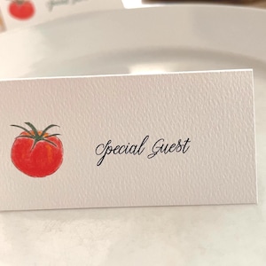 May include: A white place card with a watercolor illustration of a red tomato and green leaves. The words "Special Guest" are written in elegant script. The card is placed on a white plate.