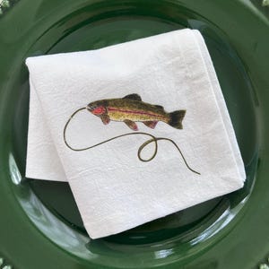 May include: A white cloth napkin featuring a detailed illustration of a rainbow trout with a fishing line trailing from its mouth. The napkin is folded and placed on top of a dark green dinner plate with a beaded edge detail along the rim.