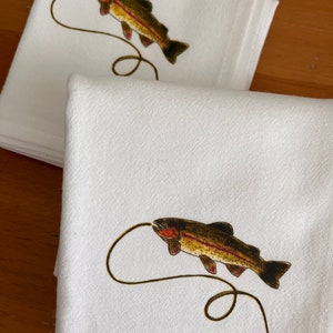 May include: Two white cotton tea towels with a brown trout fish design. The fish is depicted jumping out of the water with a fishing line attached to its mouth.