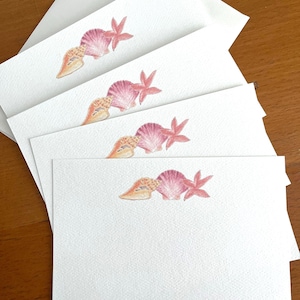 Peut inclure: Cartes de papeterie blanches avec des illustrations à l'aquarelle de coquillages et d'étoiles de mer dans des tons de rose, orange et beige. Les cartes sont empilées, avec quelques enveloppes visibles en arrière-plan.