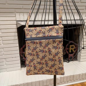 May include: A beige crossbody bag with a repeating pattern of sock monkey figures wearing red and white hats. The bag has a blue zipper and a matching strap. The bag is displayed on a black wire mannequin.