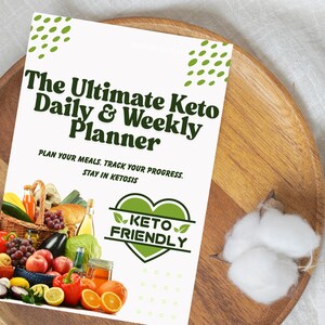 May include: A white printable planner with green text that reads "The Ultimate Keto Daily & Weekly Planner". The text "PLAN YOUR MEALS. TRACK YOUR PROGRESS. STAY IN KETOSIS" is also included. A green heart with the text "KETO FRIENDLY" is in the lower right corner. A basket of colorful fruits and vegetables is in the lower left corner.