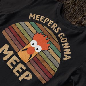 May include: Black t-shirt featuring a cartoon character with orange hair and a red nose, set against a retro rainbow design. The text "MEEPERS GONNA MEEP" is arched above and below the character.