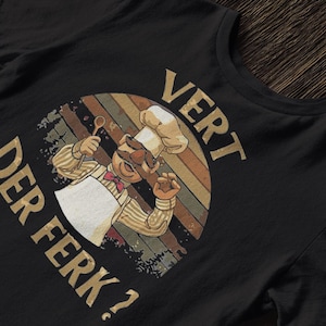 May include: Black t-shirt featuring a cartoon chef with a white apron and chef's hat, holding a spoon. The design includes the text "VERT DER FERK?" in a vintage style, with a rainbow-colored background.