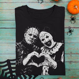 Pode incluir: Uma t-shirt preta de gola redonda com uma impressão gráfica branca de alto contraste de duas personagens de filmes de terror, Pinhead e Art the Clown, lado a lado, formando um coração com as mãos no centro da peça.