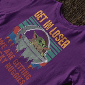 K&ouml;nnte beinhalten: Lilafarbenes T-Shirt mit einer Grafik eines Charakters in einer Kapsel und dem Text "GET IN LOSER WE ARE GETTING CHICKY NUGGIES" in Orange und Wei&szlig;. Das Design enth&auml;lt einen gr&uuml;n, blau und rot gestreiften Hintergrund.