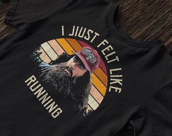 Puede incluir: Camiseta negra con un gráfico de un hombre con barba larga y gorra, sobre un arco de colores de atardecer. El texto dice "I JUST FELT LIKE RUNNING."