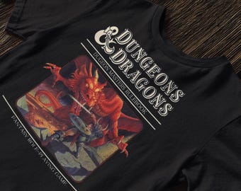 Puede incluir: Camiseta negra con una ilustración de fantasía de un dragón rojo luchando contra un caballero. La camiseta incluye el texto "DUNGEONS & DRAGONS" y "Dungeon Masters Rulebook" en blanco, junto con "Fantasy Role-Playing Game".