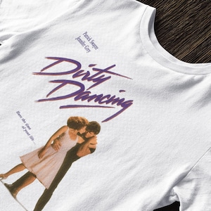 May include: White t-shirt with the film title "Dirty Dancing" in purple script. The shirt features an image of the film's stars and the phrase "Have the time of your life..." printed below the title.
