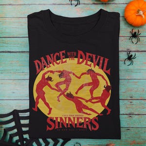 Puede incluir: Una camiseta negra de cuello redondo con un estampado gráfico de cinco figuras rojas con cuernos bailando en círculo frente a una luna amarilla. El texto dice DANCE WITH THE DEVIL SINNERS en letras rojas, con texto más pequeño debajo.
