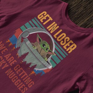 K&ouml;nnte beinhalten: Burgunderfarbenes T-Shirt mit einer Grafik von Grogu aus The Mandalorian in einer Kapsel, mit dem Text "GET IN LOSER WE ARE GETTING CHICKY NUGGIES". Das Design beinhaltet ein Retro-Farbschema und Waldmotive.