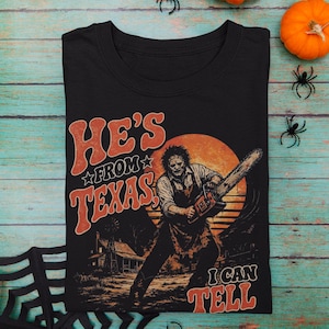 Pode incluir: Uma t-shirt preta com uma estampa gráfica de uma personagem segurando uma motosserra em frente a uma grande lua laranja. O texto "HE'S FROM TEXAS" e "I CAN TELL" está escrito numa fonte laranja retro. O design inclui uma casa.