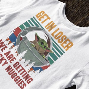 K&ouml;nnte beinhalten: Wei&szlig;es T-Shirt mit einer Grafik eines gr&uuml;nen Alien-Charakters in einer Kapsel und dem Text "GET IN LOSER WE ARE GETTING CHICKY NUGGIES" in Orange und Rot. Das Design hat einen Retro-Stil mit gr&uuml;nen und blauen Streifen.