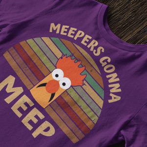 Peut inclure: T-shirt violet avec un graphisme de Beaker des Muppets. Le design comprend le texte "MEEPERS GONNA MEEP" dans un style r&eacute;tro, avec un fond ray&eacute; color&eacute; derri&egrave;re Beaker.