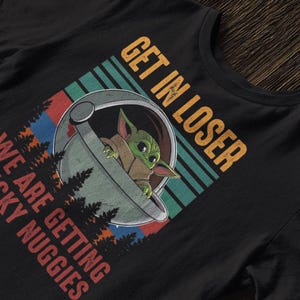 K&ouml;nnte beinhalten: Schwarzes T-Shirt mit einem Grafikdesign eines gr&uuml;nen Alien-Charakters in einer Kapsel und dem Text "GET IN LOSER WE ARE GETTING CHICKY NUGGIES" in Orange und Rot. Das Design hat einen Retro-Look.