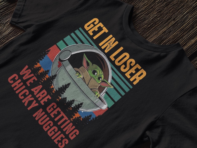 K&ouml;nnte beinhalten: Schwarzes T-Shirt mit einem Grafikdesign eines gr&uuml;nen Alien-Charakters in einer Kapsel und dem Text "GET IN LOSER WE ARE GETTING CHICKY NUGGIES" in Orange und Rot. Das Design hat einen Retro-Look.