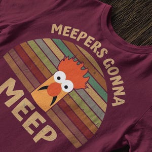 Peut inclure: T-shirt bordeaux avec un design r&eacute;tro, avec le texte "MEEPERS GONNA MEEP". Le motif comprend un personnage de dessin anim&eacute; avec des cheveux orange et un nez rond orange, sur un fond arc-en-ciel ray&eacute;. Un article de v&ecirc;tement d&eacute;contract&eacute;.
