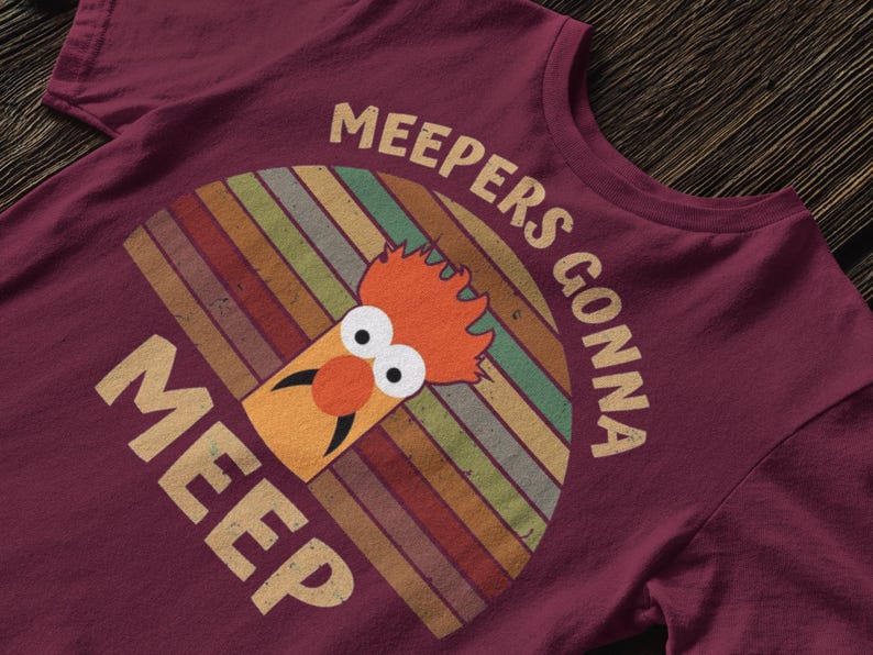 Peut inclure: T-shirt bordeaux avec un design r&eacute;tro, avec le texte "MEEPERS GONNA MEEP". Le motif comprend un personnage de dessin anim&eacute; avec des cheveux orange et un nez rond orange, sur un fond arc-en-ciel ray&eacute;. Un article de v&ecirc;tement d&eacute;contract&eacute;.