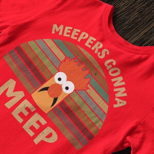 Peut inclure: T-shirt rouge avec un personnage de dessin anim&eacute; et le texte "MEEPERS GONNA MEEP". Le motif est de style r&eacute;tro avec un fond ray&eacute; color&eacute; et une illustration de personnage fantaisiste.