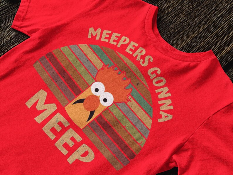 Peut inclure: T-shirt rouge avec un personnage de dessin anim&eacute; et le texte "MEEPERS GONNA MEEP". Le motif est de style r&eacute;tro avec un fond ray&eacute; color&eacute; et une illustration de personnage fantaisiste.