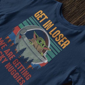 K&ouml;nnte beinhalten: Marineblaues T-Shirt mit einer Grafik von Grogu in einer Kapsel und dem Text "GET IN LOSER WE ARE GETTING CHICKY NUGGIES". Das Design enth&auml;lt Wald- und Streifenelemente.