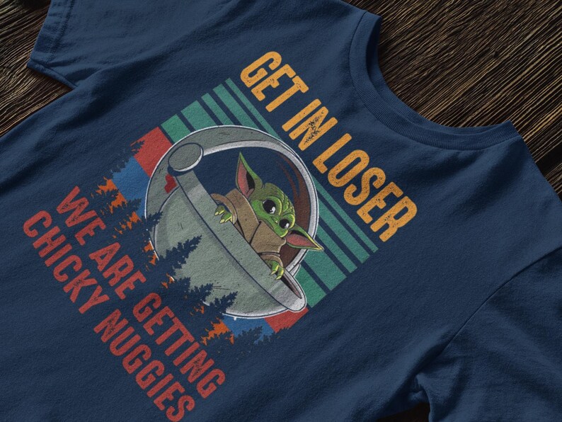 K&ouml;nnte beinhalten: Marineblaues T-Shirt mit einer Grafik von Grogu in einer Kapsel und dem Text "GET IN LOSER WE ARE GETTING CHICKY NUGGIES". Das Design enth&auml;lt Wald- und Streifenelemente.