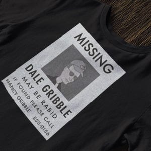 Può includere: T-shirt nera con un design di poster "Missing" invecchiato. Il poster include un'immagine in scala di grigi di un uomo con berretto e occhiali da sole, con il nome "Dale Gribble" e il testo "May be rabid."