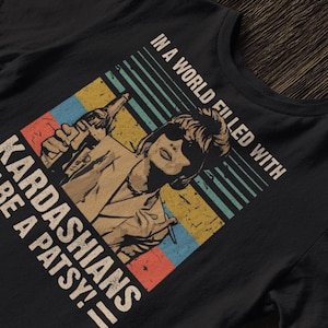 以下が含まれることがあります： レトロスタイルのグラフィックが特徴の黒いTシャツ。「IN A WORLD FILLED WITH KARDASHIANS BE A PATSY!」のテキストと、ボトルを持つ女性のイラストが、カラフルな背景に描かれています。