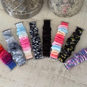 May include: A collection of scrunchie-style watch bands in various colors and patterns. The bands include a rainbow gradient, tie-dye, floral, and solid black. The bands are made of fabric and have silver connectors.