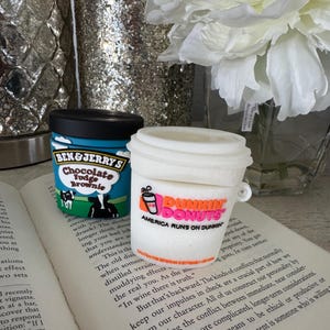 May include: Two miniature food-themed items: a Ben & Jerry's Chocolate Fudge Brownie ice cream container and a Dunkin' Donuts coffee cup. The Dunkin' Donuts cup has the text "AMERICA RUNS ON DUNKIN". Both items are on an open book.