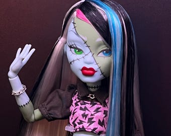 May include: A doll with green skin, black and white hair with blue and pink streaks, and red lips. The doll is wearing a black top with a pink lightning bolt design, black pants, and a silver chain belt. The doll has stitches on its face and arms.