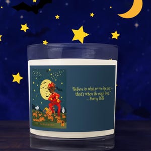 May include: A clear glass candle with a dark blue label featuring a whimsical illustration of a cat in a red costume in a pumpkin patch under a full moon. The candle has the quote, "Believe in what no one else sees - that's where the magic lives."