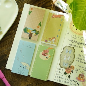 Peut inclure: Plusieurs blocs-notes colorés avec des illustrations, dont un chat sous un parapluie, un chat jouant avec une pelote de laine et un ours dans un paysage. Des tubes d'aquarelle et un stylo rose sont également visibles.