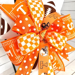 May include: A decorative bow featuring orange and white ribbons with football and checkerboard patterns. The bow includes the letter "T" and football field markings. Accented with white polka dots and a football graphic.