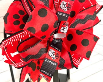 Puede incluir: Una linterna negra con un lazo rojo y negro. El lazo está hecho de cinta con un patrón de lunares y un patrón repetido de un bulldog con las palabras "Georgia Bulldogs".
