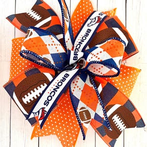 May include: A large orange, white, and blue bow with a football and "Broncos" design. The bow is made of ribbon and has a polka dot pattern.