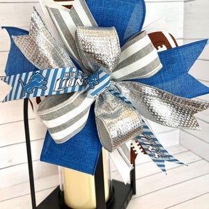 May include: A black lantern with a blue, white, and silver bow. The bow has a ribbon with the words "Detroit Lions" on it. The lantern is decorated with footballs.