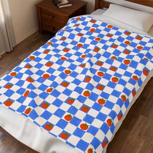 Puede incluir: Una manta a cuadros azules y blancos con un patrón repetido de tomates naranjas. La manta está extendida sobre una cama, mostrando los colores vibrantes y el diseño lúdico. La manta está hecha de un material suave.