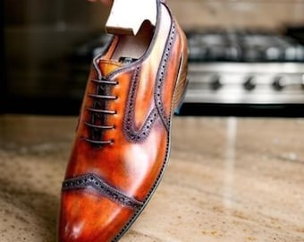 May include: A close-up of a brown leather oxford shoe with a brogue design. The shoe has a lace-up closure and a pointed toe.