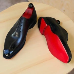 May include: A pair of black leather dress shoes with red soles and interior lining. The shoes are displayed on a light wooden surface. The shoes have a classic lace-up design and a glossy finish.
