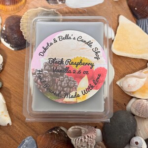 Może przedstawiać: Kwadratowy wosk do topienia w przezroczystym plastikowym pojemniku. Wosk jest szary i ma etykietę z napisem "Dakota & Bella's Candle Shop" i "Black Raspberry Vanilla 2.6 oz". Etykieta zawiera również zdjęcie czarnych malin i wanilii.